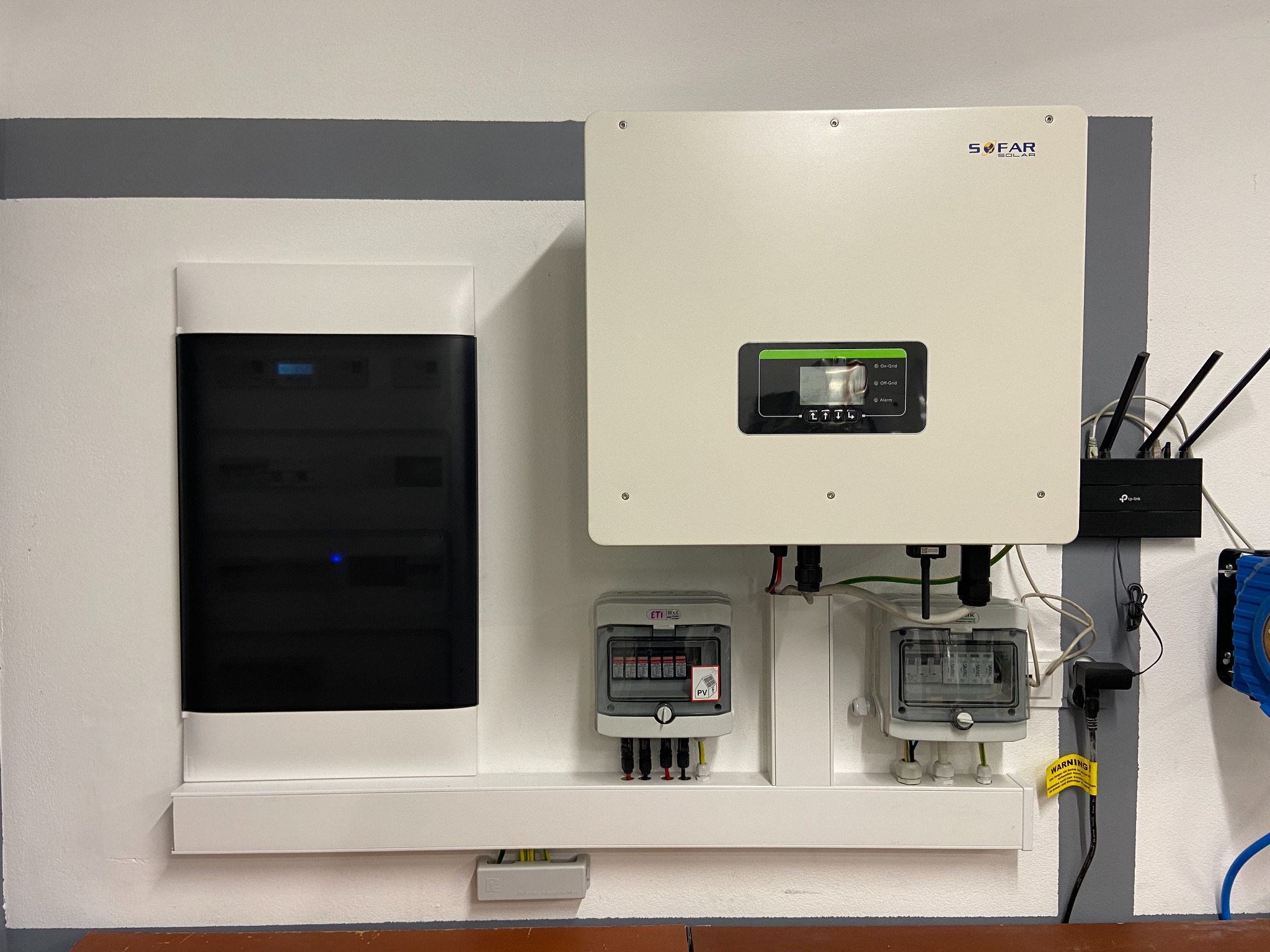Zestawienie urządzeń: rozdzielnica elektryczna, falownik SOFAR Solar z wyświetlaczem i router TP-Link na ścianie z szarą listwą dekoracyjną.