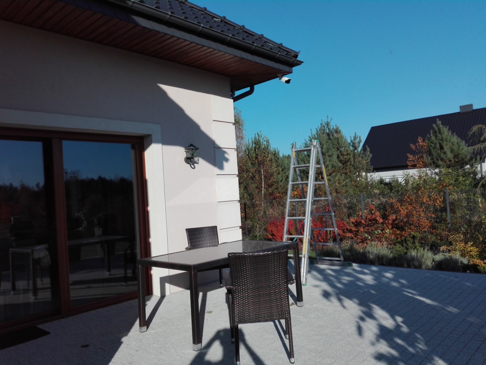 Kamera monitoringu zamontowana na elewacji domu, widoczna drabina oparta o ścianę, stół i krzesła na tarasie w słoneczny dzień.