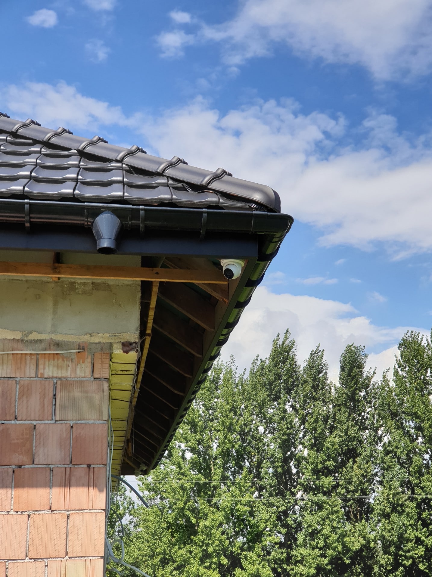 Kamera kopułkowa zamontowana pod okapem dachu, widok z dołu na tle błękitnego nieba i zielonych drzew, widoczne elementy konstrukcyjne budynku w trakcie budowy.