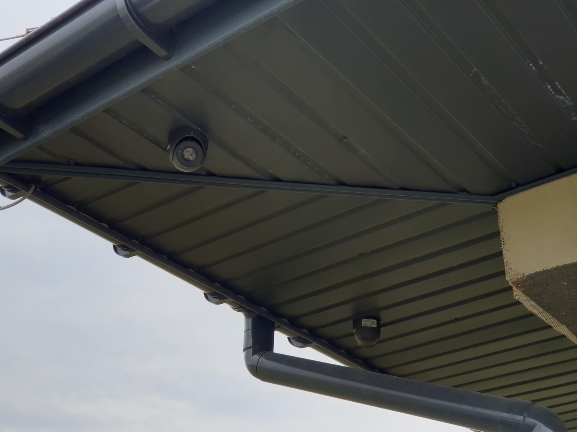 Dwie kamery kopułkowe zamontowane pod metalowym zadaszeniem, widoczne rynny i fragment elewacji budynku.