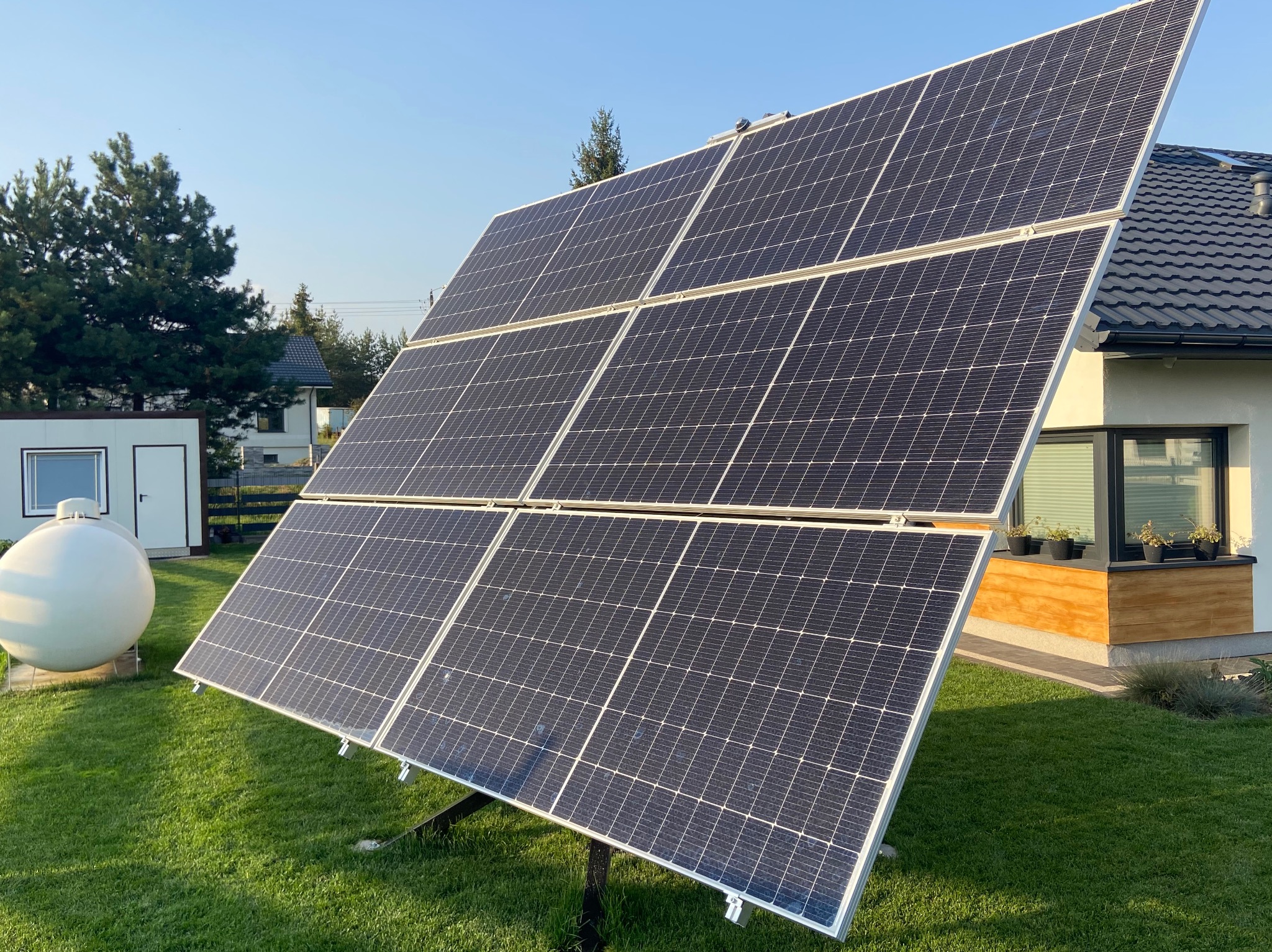 Panel fotowoltaiczny na trawniku przed domem jednorodzinnym, widoczny zbiornik na gaz i fragment elewacji z oknem.