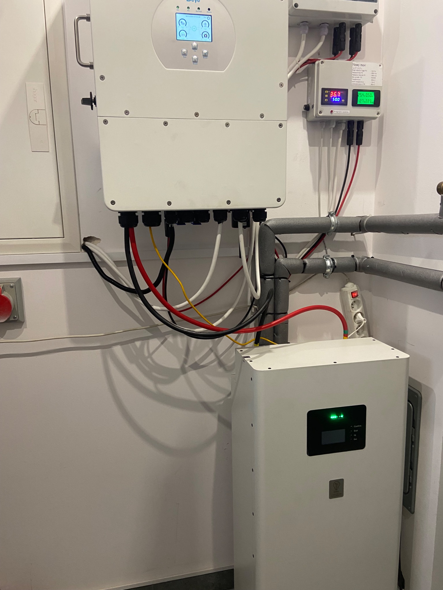 Biała bateria magazynu energii z podłączonymi przewodami, obok inwertera fotowoltaicznego i dodatkowego modułu pomiarowego na białej ścianie w pomieszczeniu technicznym.