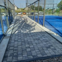 Rzepka-Usługi brukarskie - Nowoczesna nawierzchnia z szarej kostki brukowej przy błękitnym korcie tenisowym, wykończona drobnym żwirem i metalową siatką. Dwie drewniane ławki w tle.