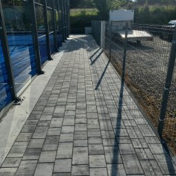 Rzepka-Usługi brukarskie - Szara kostka brukowa ułożona wzdłuż metalowego ogrodzenia i kortu do padla. Cień rzucany przez słupki ogrodzenia. Trawa i drzewa w tle.