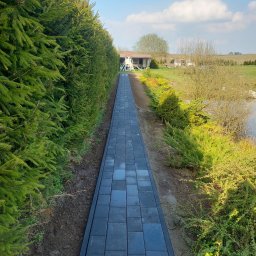 Rzepka-Usługi brukarskie - Ścieżka z ciemnej kostki brukowej prowadząca przez ogród w Kościerzynie, otoczona tujami i krzewami, w tle dom i błękitne niebo.