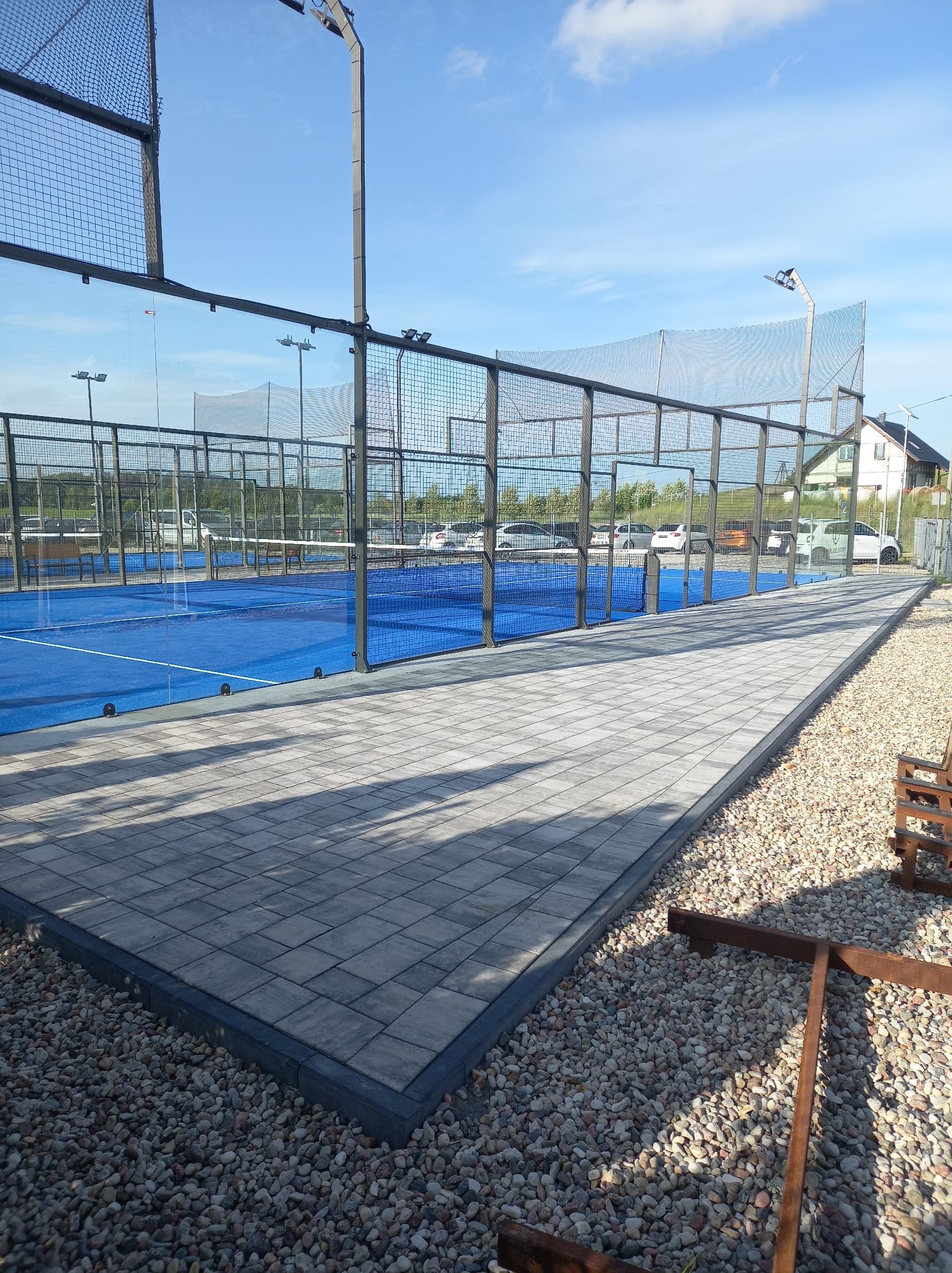 Nowoczesny kort do padla z niebieską nawierzchnią i szarą kostką brukową wokół, otoczony kamieniami i metalową siatką, w słoneczny dzień.