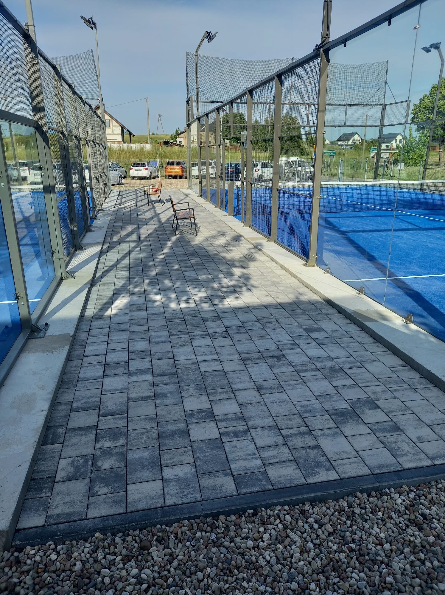 Nowoczesna nawierzchnia z szarej kostki brukowej przy błękitnym korcie tenisowym, wykończona drobnym żwirem i metalową siatką. Dwie drewniane ławki w tle.