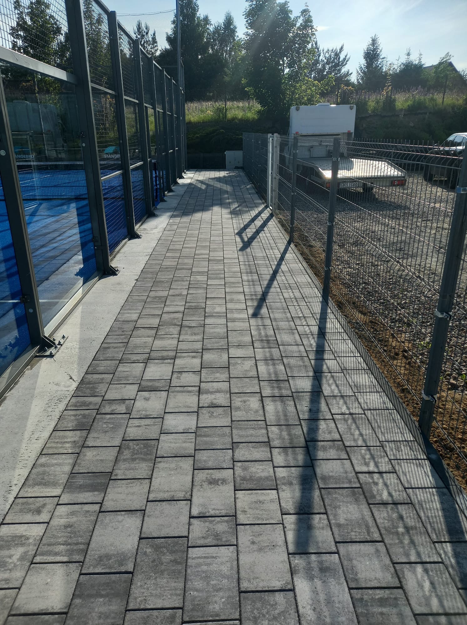 Szara kostka brukowa ułożona wzdłuż metalowego ogrodzenia i kortu do padla. Cień rzucany przez słupki ogrodzenia. Trawa i drzewa w tle.