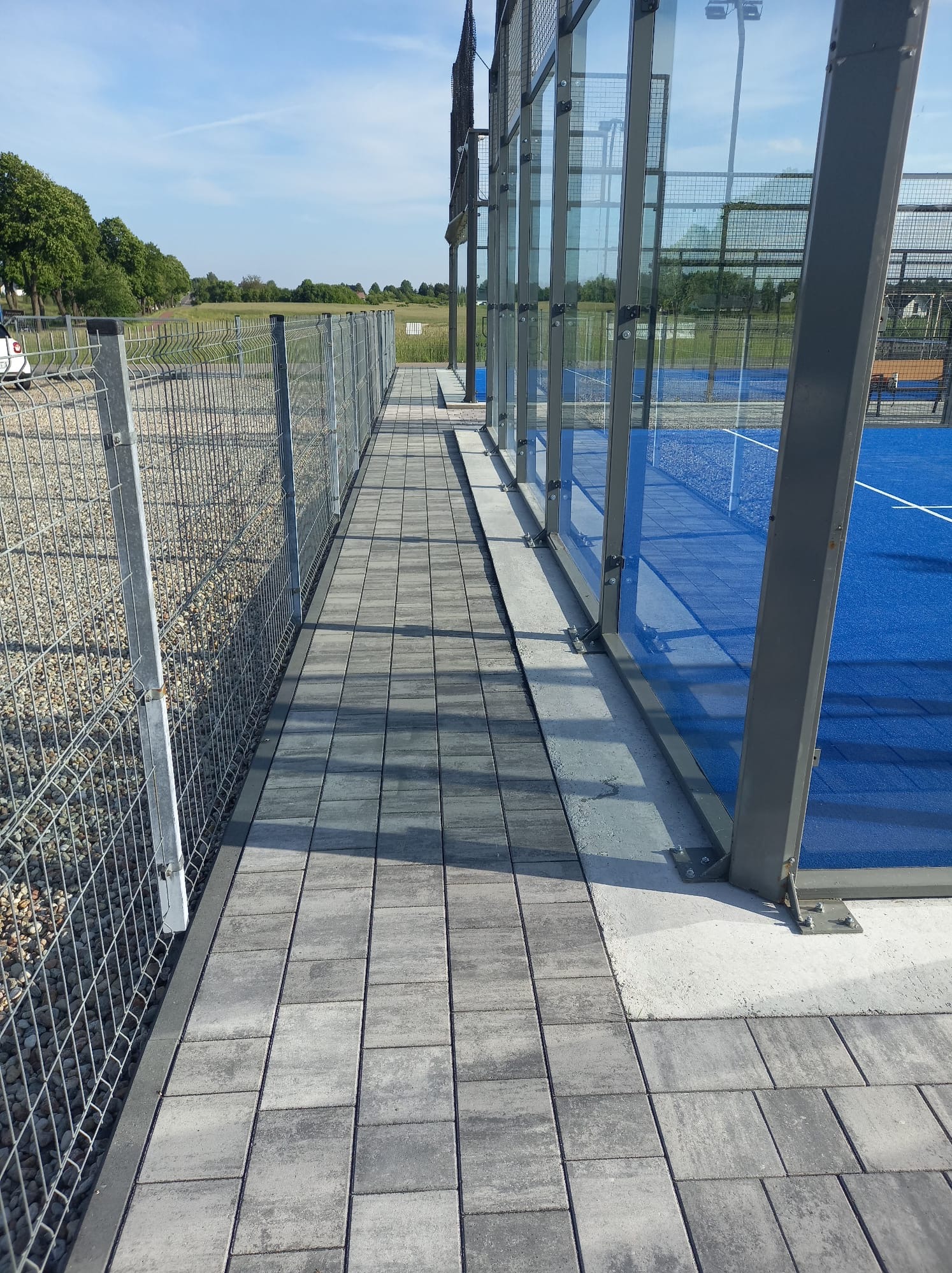 Szara kostka brukowa ułożona wzdłuż ogrodzenia i szklanej ściany kortu tenisowego. Widok na zielone pola i błękitne niebo w tle.