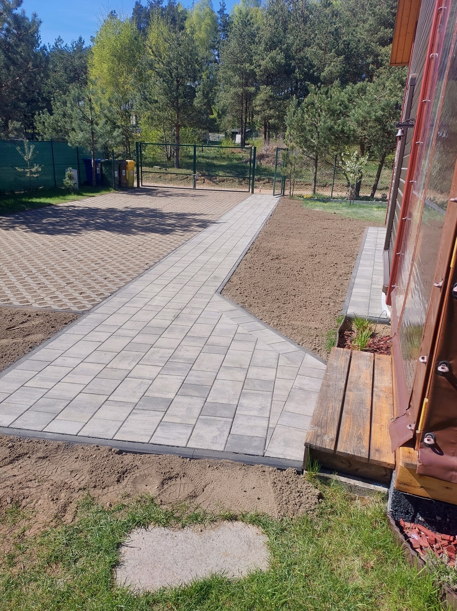 Ścieżka z jasnoszarej kostki brukowej prowadzi przez ogród, łącząc budynek z bramą w tle. Część powierzchni wyłożona ażurowymi płytami.