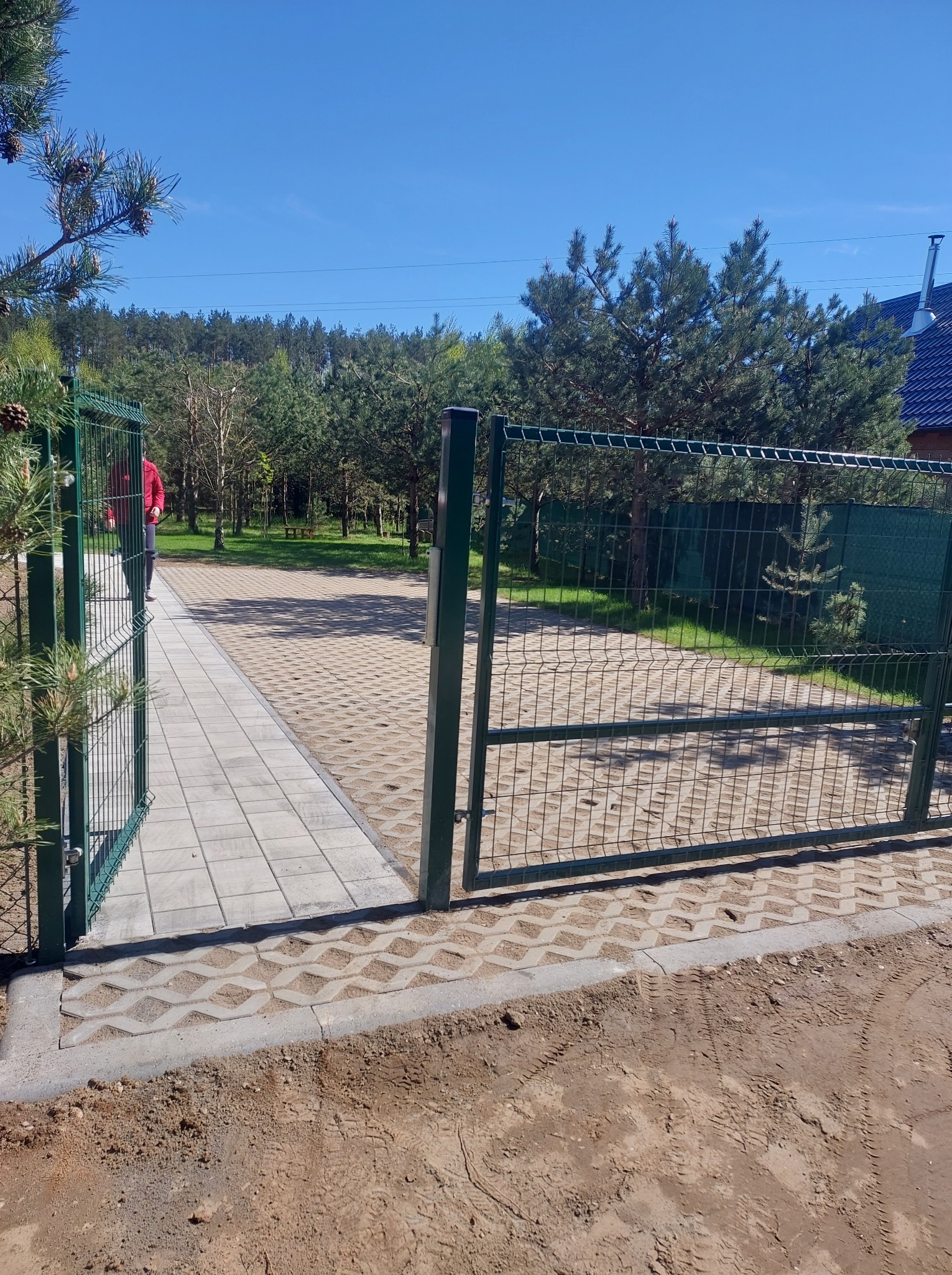 Otwarta, zielona, metalowa furtka z siatki. Za nią ścieżka z jasnych płyt i ażurowa kostka brukowa. W tle drzewa i budynek.