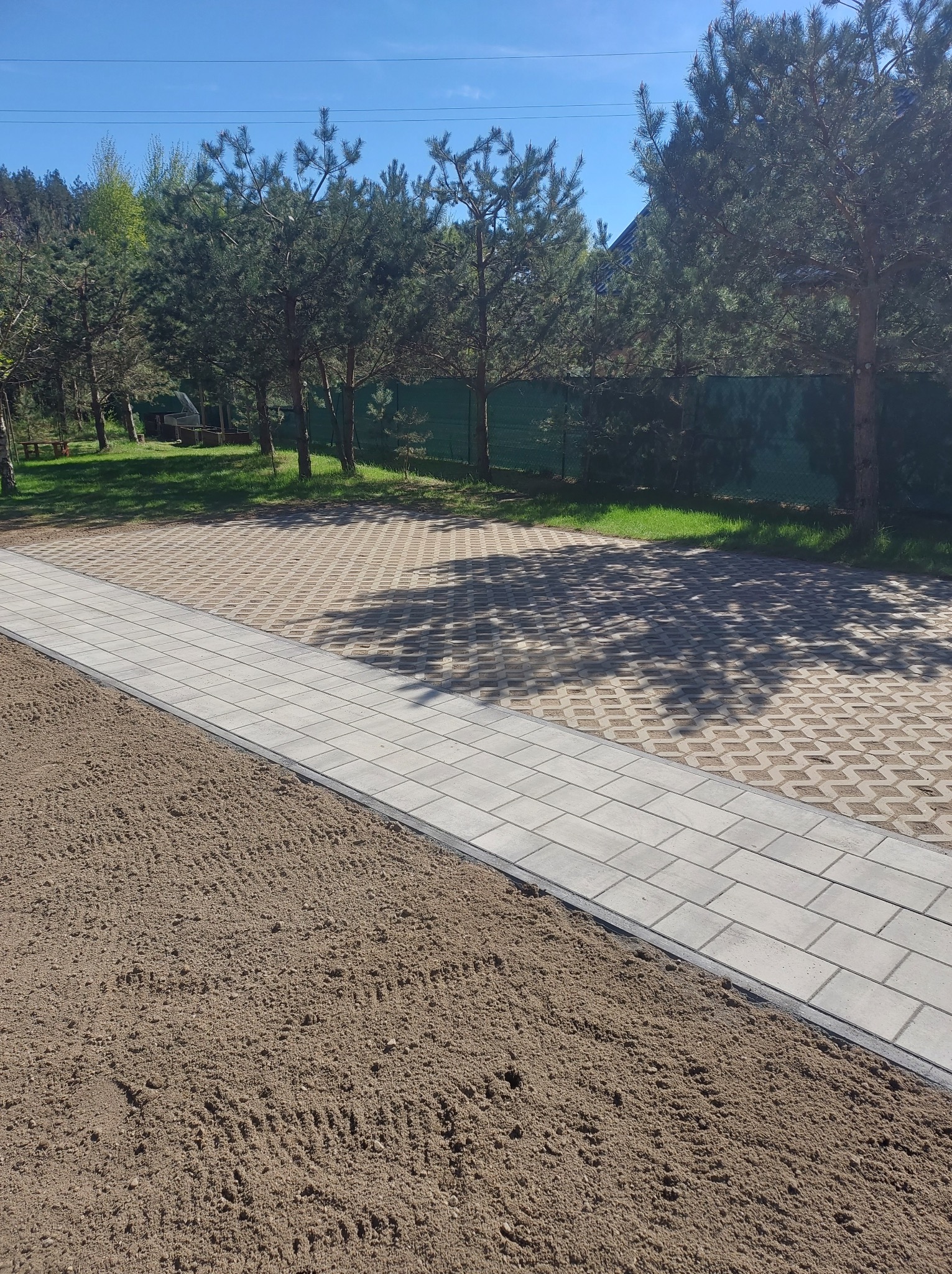 Ścieżka z jasnoszarej kostki brukowej oddziela przygotowany teren pod budowę od ażurowej nawierzchni parkingu. W tle drzewa i zielone ogrodzenie.