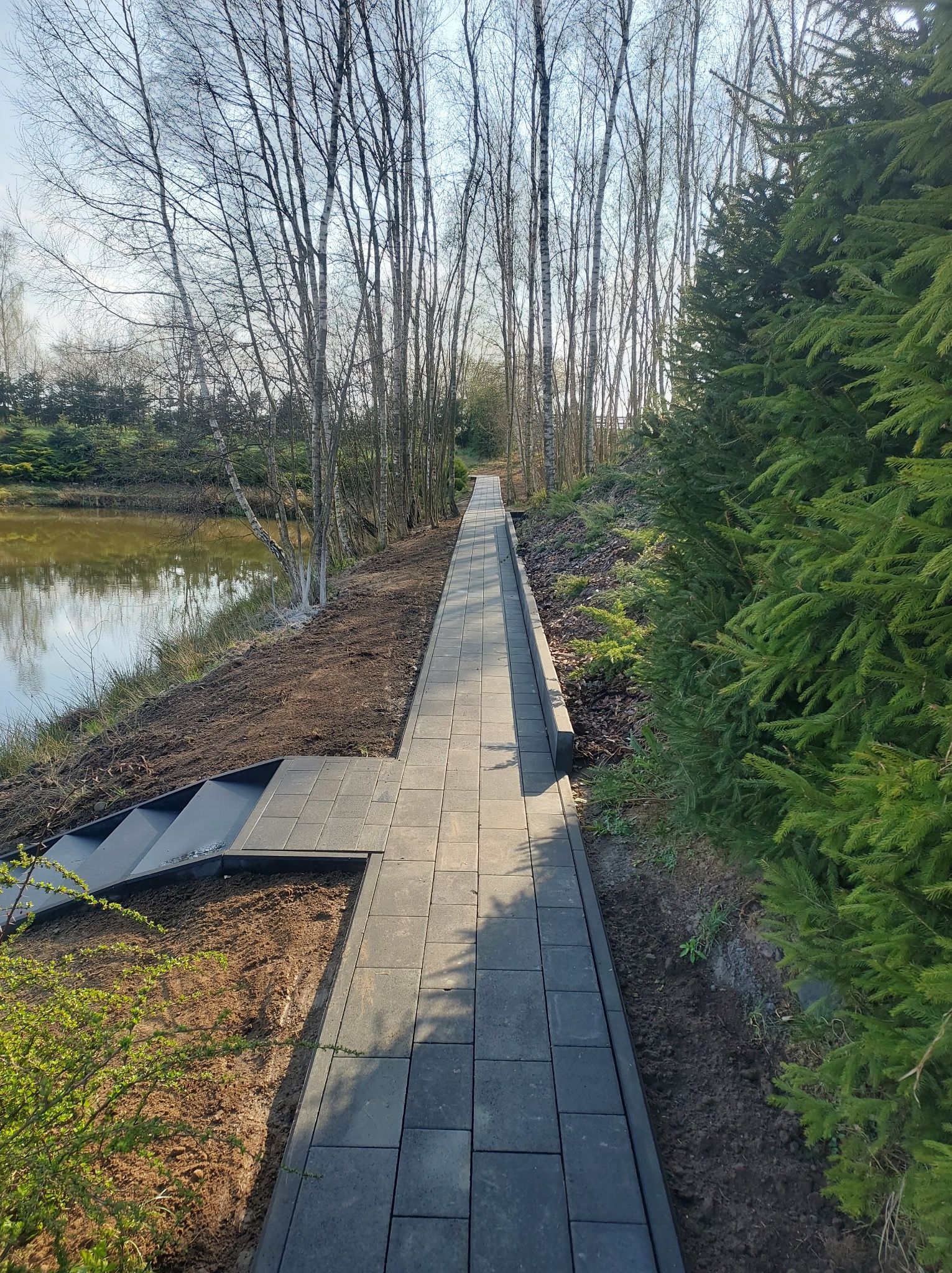 Ścieżka z kostki brukowej prowadząca wzdłuż jeziora, zakończona schodami. Po prawej stronie rosną gęste, zielone drzewa iglaste, po lewej widać brzeg jeziora.