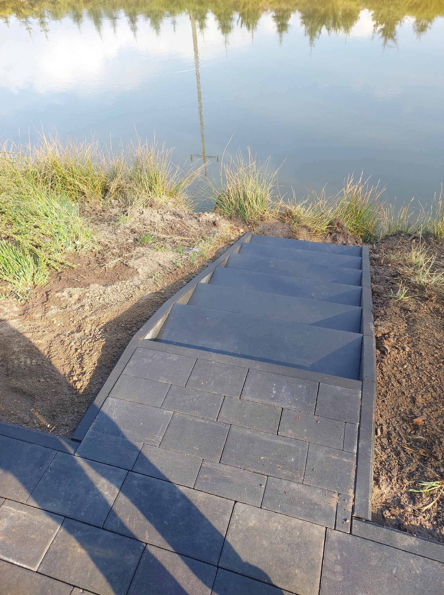 Nowoczesne, grafitowe schody z kostki brukowej prowadzące do jeziora, otoczone trawą i ziemią. Minimalistyczny design, stonowana kolorystyka.