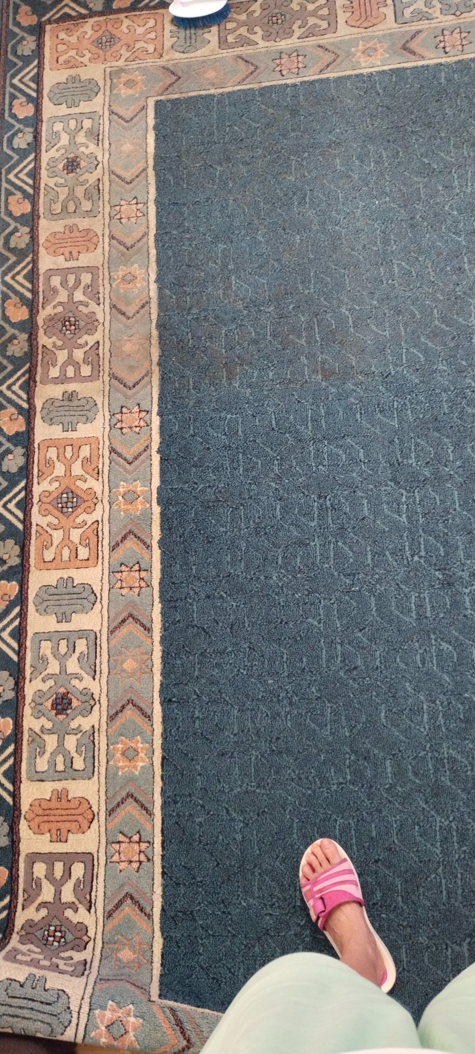 Fragment niebieskiego dywanu z orientalnym wzorem obok stopy w różowym klapku, widoczne ślady czyszczenia na powierzchni.