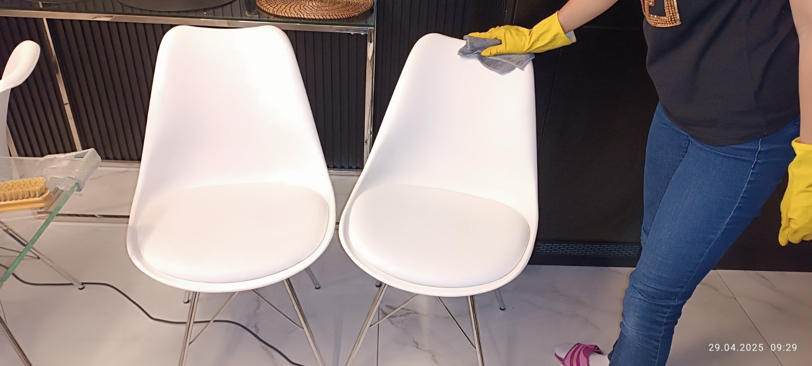 Osoba w żółtych rękawicach czyści białe krzesło z szarą ściereczką. W tle widoczne inne meble i elementy wyposażenia wnętrza.