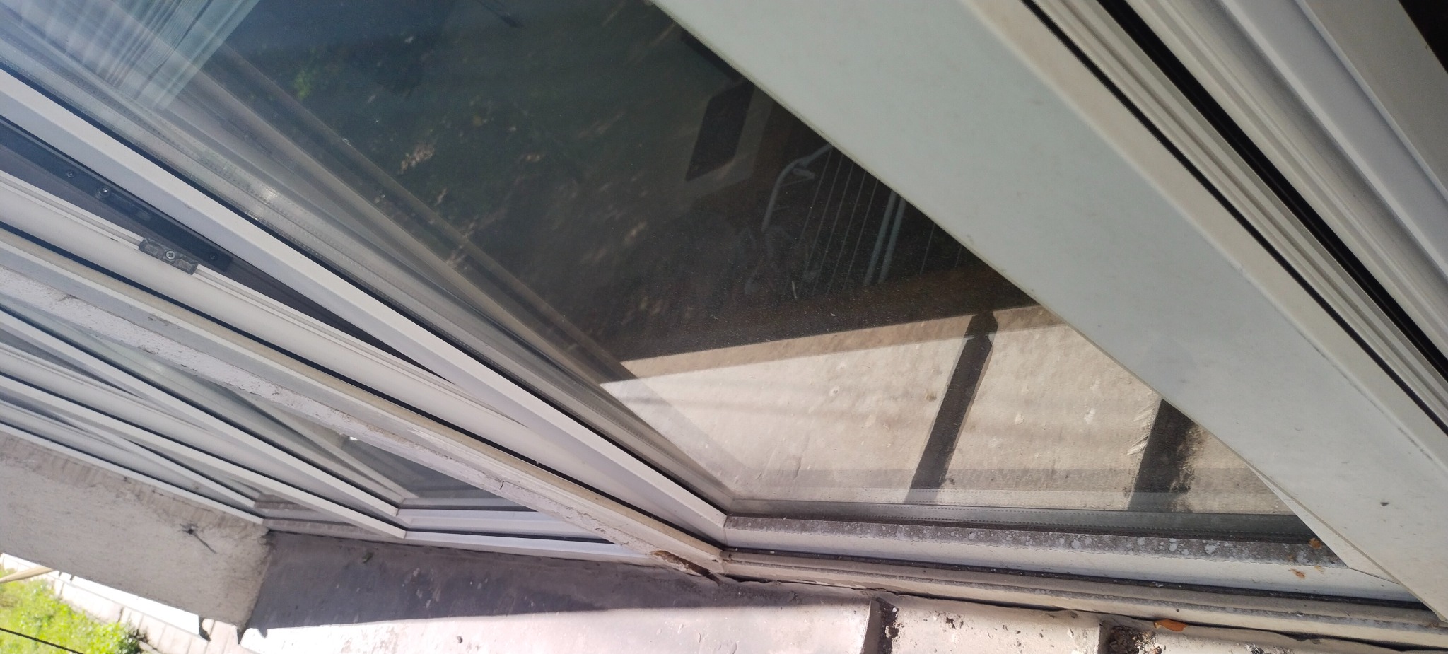 Brudne okno z białymi ramami, widoczne zabrudzenia na szybie i wzdłuż ramy. Perspektywa z dołu, fragment ściany i zieleni w tle.