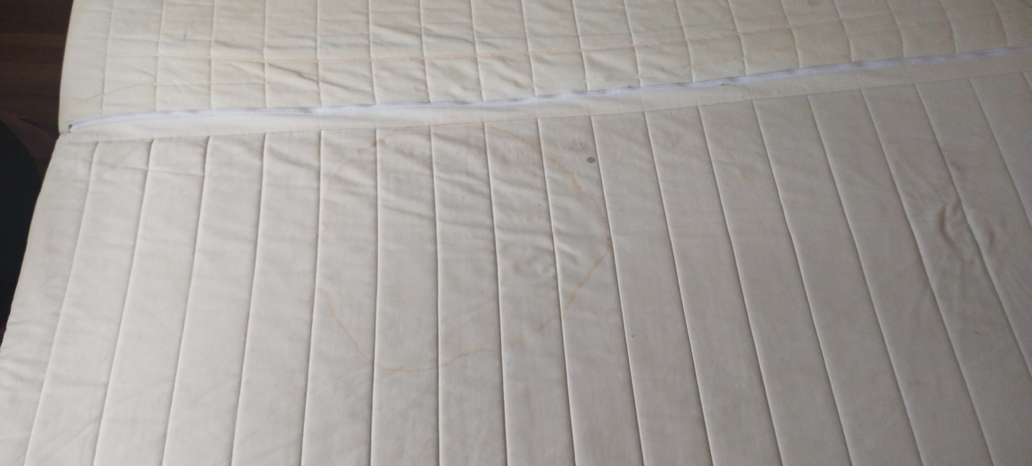 Materac z beżową tapicerką, widoczne plamy i zamek błyskawiczny. Powierzchnia materaca podzielona na podłużne pasy.