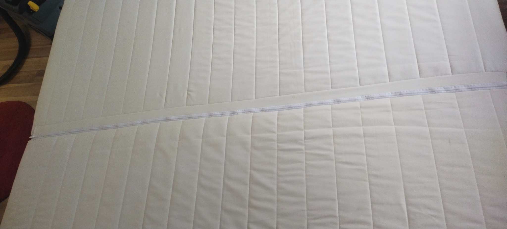 Jasny materac z zamkiem błyskawicznym, widoczny od góry, z delikatnym wzorem pikowania. W tle fragment odkurzacza i czerwony element.