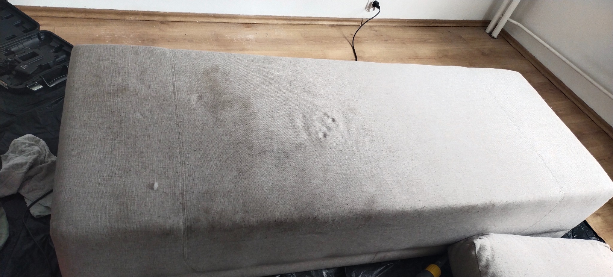 Beżowa, zabrudzona sofa przed praniem, widoczne plamy i ślady użytkowania na tapicerce. Obok leżą narzędzia do czyszczenia.