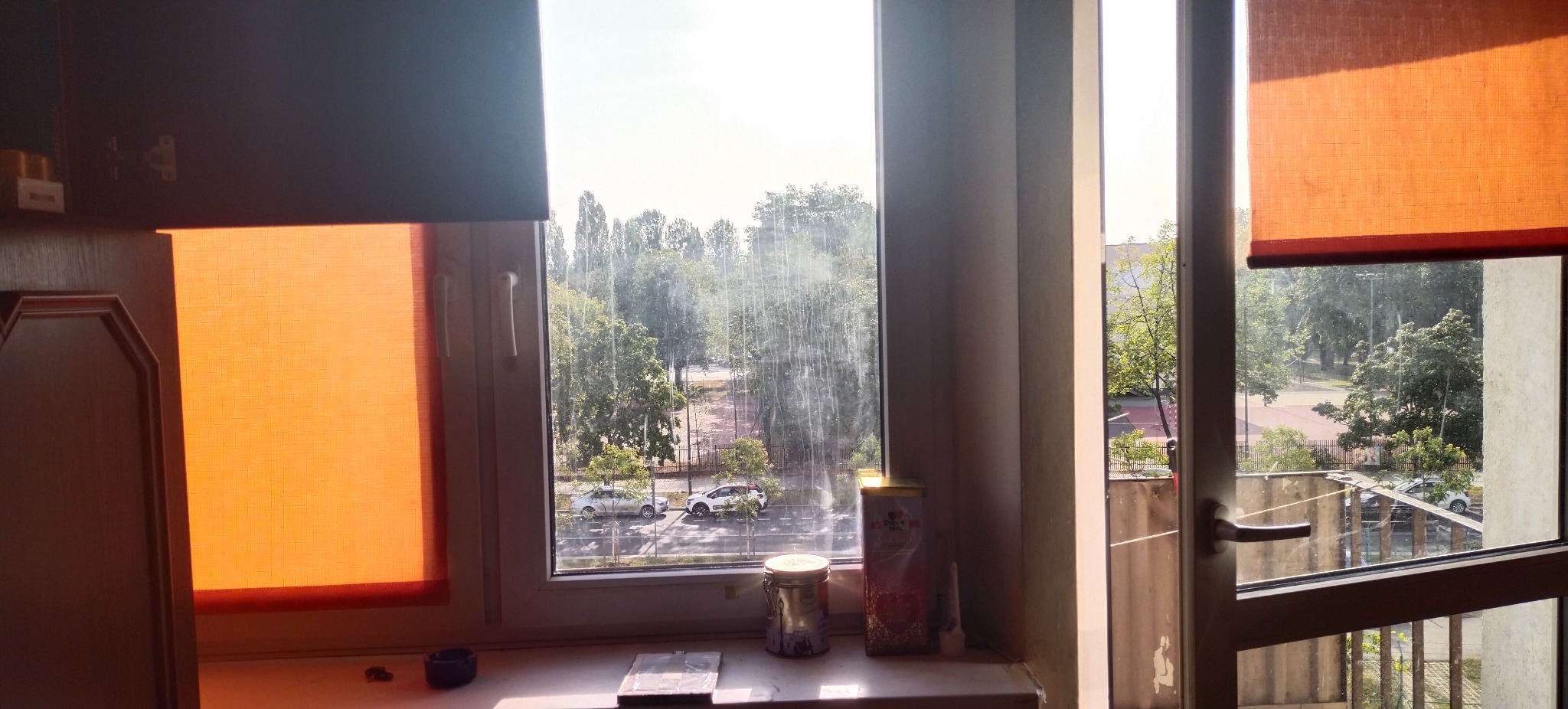 Widok z okna na zieleń miejską, z widocznymi smugami brudu na szybie i pomarańczowymi roletami po bokach. Na parapecie stoją puszki i drobne przedmioty.