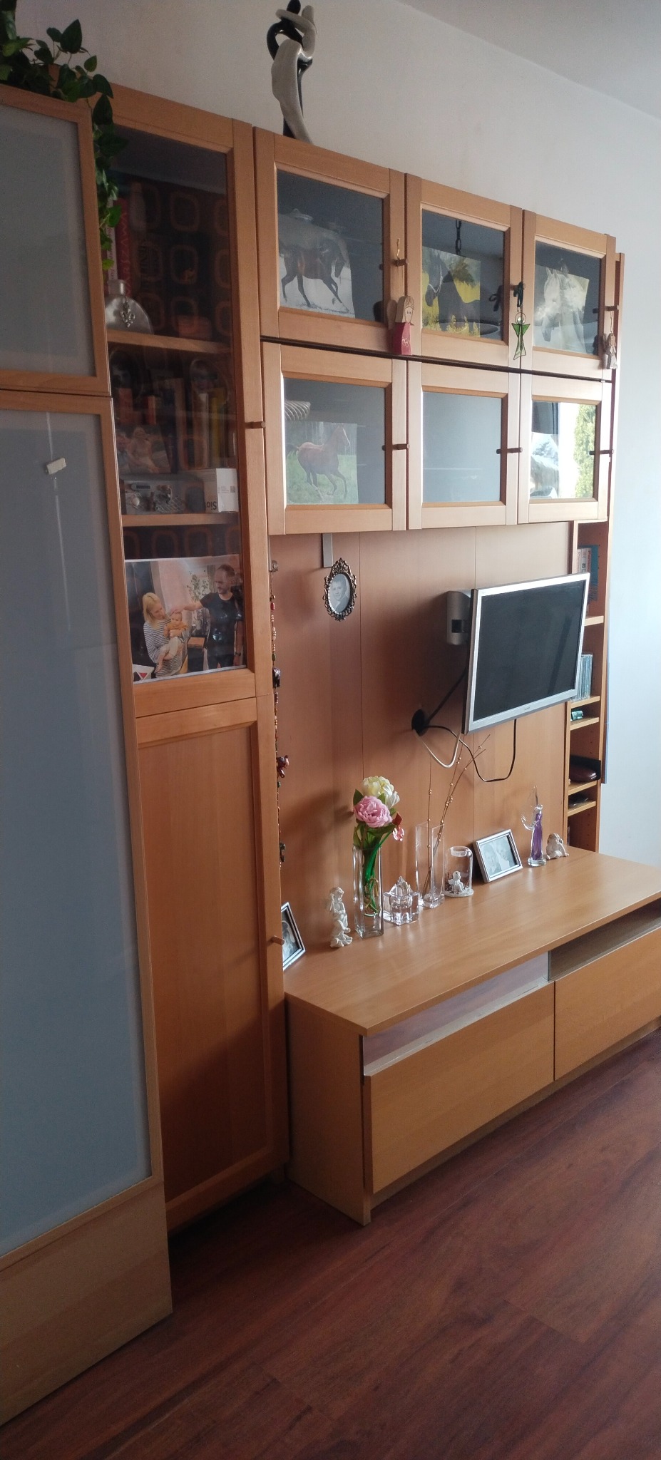 Jasne drewniane meble w salonie z telewizorem, kwiatami i dekoracjami. Ustawienie tworzy przytulną i uporządkowaną przestrzeń.