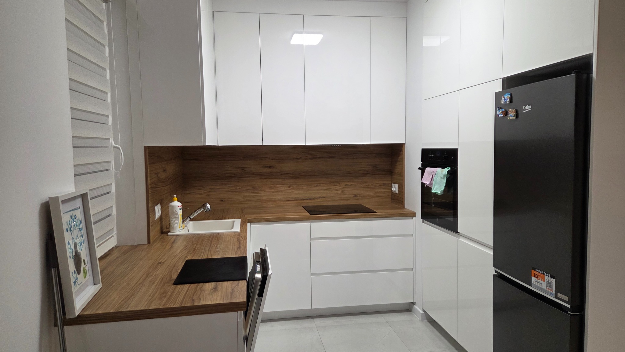 Nowoczesna, jasna kuchnia z białymi szafkami i drewnianym blatem. Widoczny zlew, płyta indukcyjna, piekarnik i czarna lodówka. Minimalistyczny design, szare płytki na podłodze.