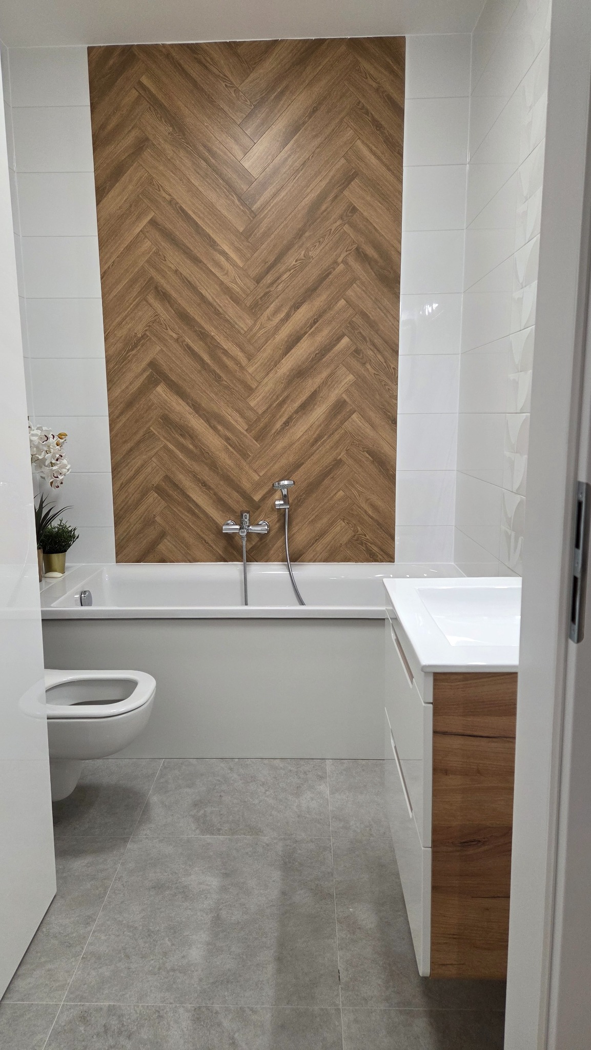 Nowoczesna łazienka po remoncie: białe płytki, drewniana mozaika nad wanną, szare płytki podłogowe, umywalka z drewnianymi elementami. Widok zza otwartych drzwi.