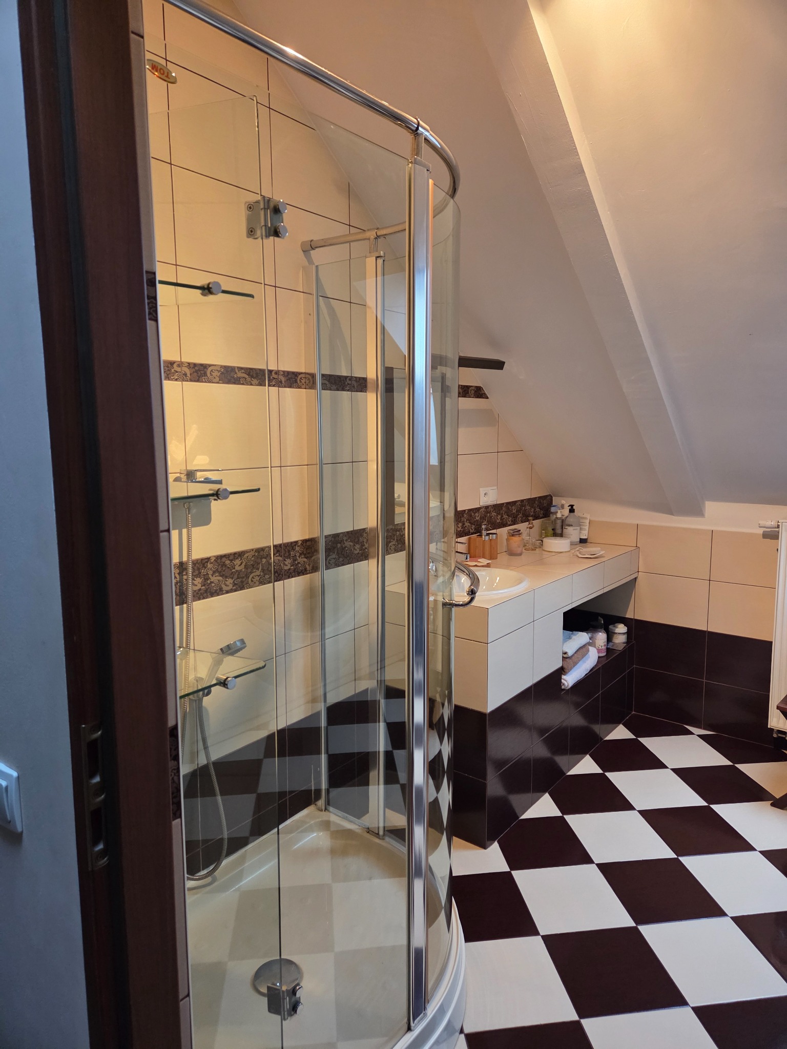 Ukończona łazienka na poddaszu z kabiną prysznicową, umywalką i czarno-białą podłogą w szachownicę. Widoczne płytki i akcesoria łazienkowe.