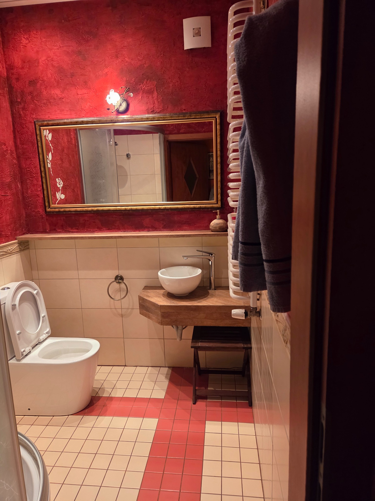 Czerwona łazienka po remoncie: biała ceramika, drewniany blat z umywalką, lustro w złotej ramie. Widoczne płytki podłogowe w odcieniach czerwieni i beżu.