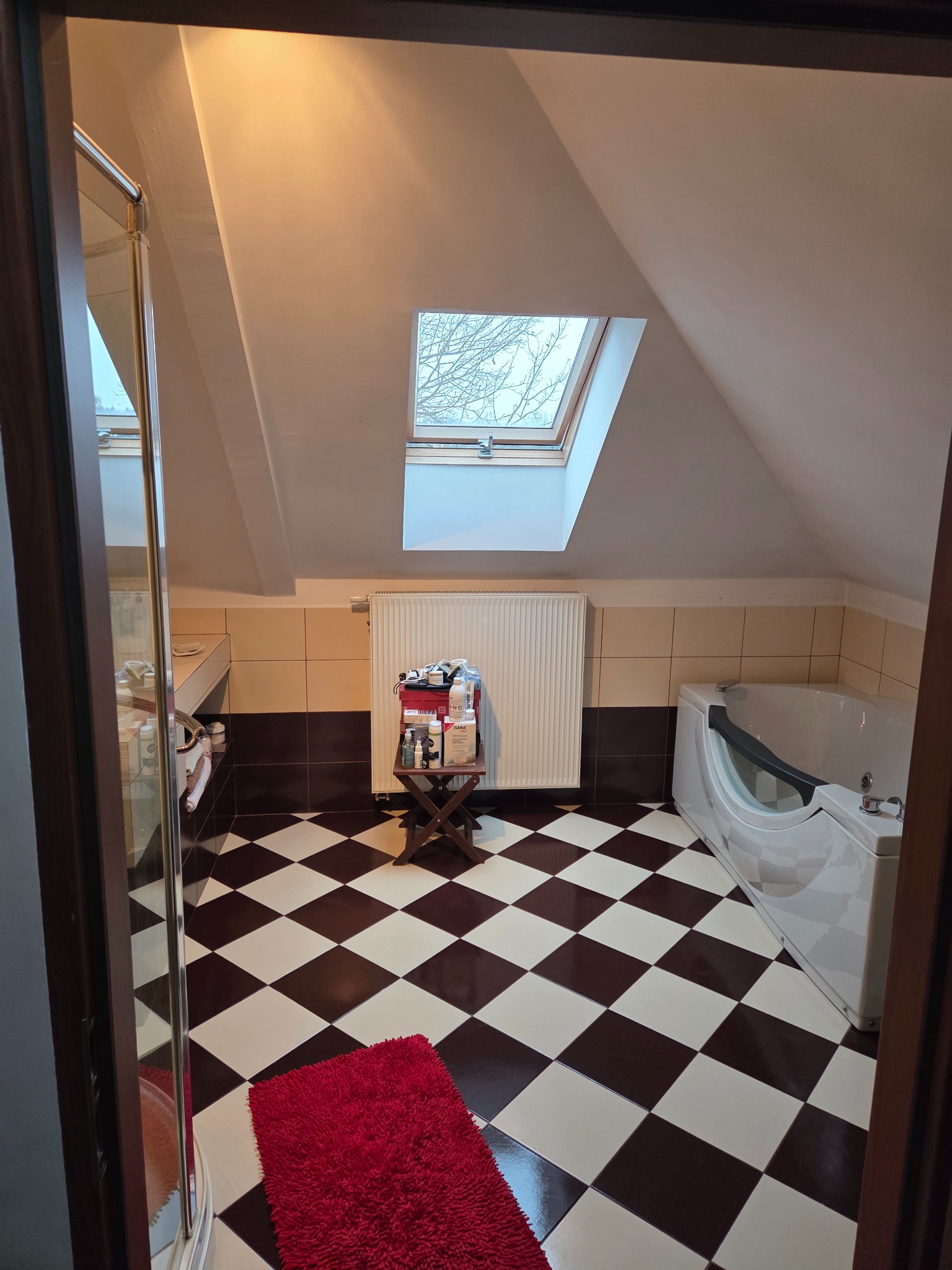 Umywalka, wanna i szachownica na podłodze w łazience na poddaszu z oknem dachowym. Na taborecie kosmetyki i środki czystości. Widok zza framugi drzwi.