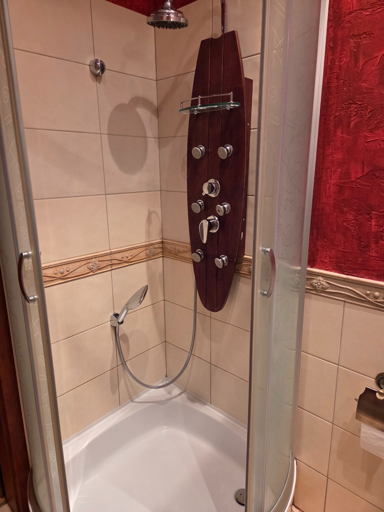 Wnętrze kabiny prysznicowej z panelem sterującym z drewna, chromowaną słuchawką i deszczownicą. Beżowe płytki i czerwona ściana w tle. Widok z otwartych drzwi kabiny.