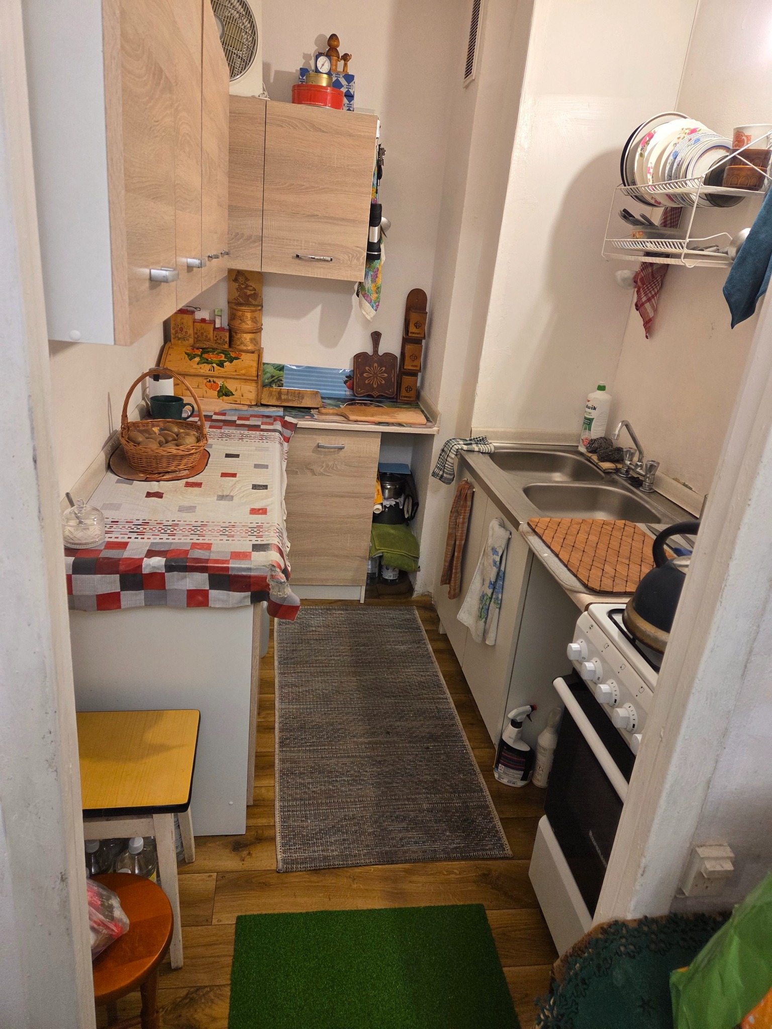 Wnętrze małej kuchni z drewnianymi szafkami, metalowym zlewem i kuchenką. Na blacie koszyk z owocami i tekstylia w kratę. Widoczny dywanik i butelki pod stołem.