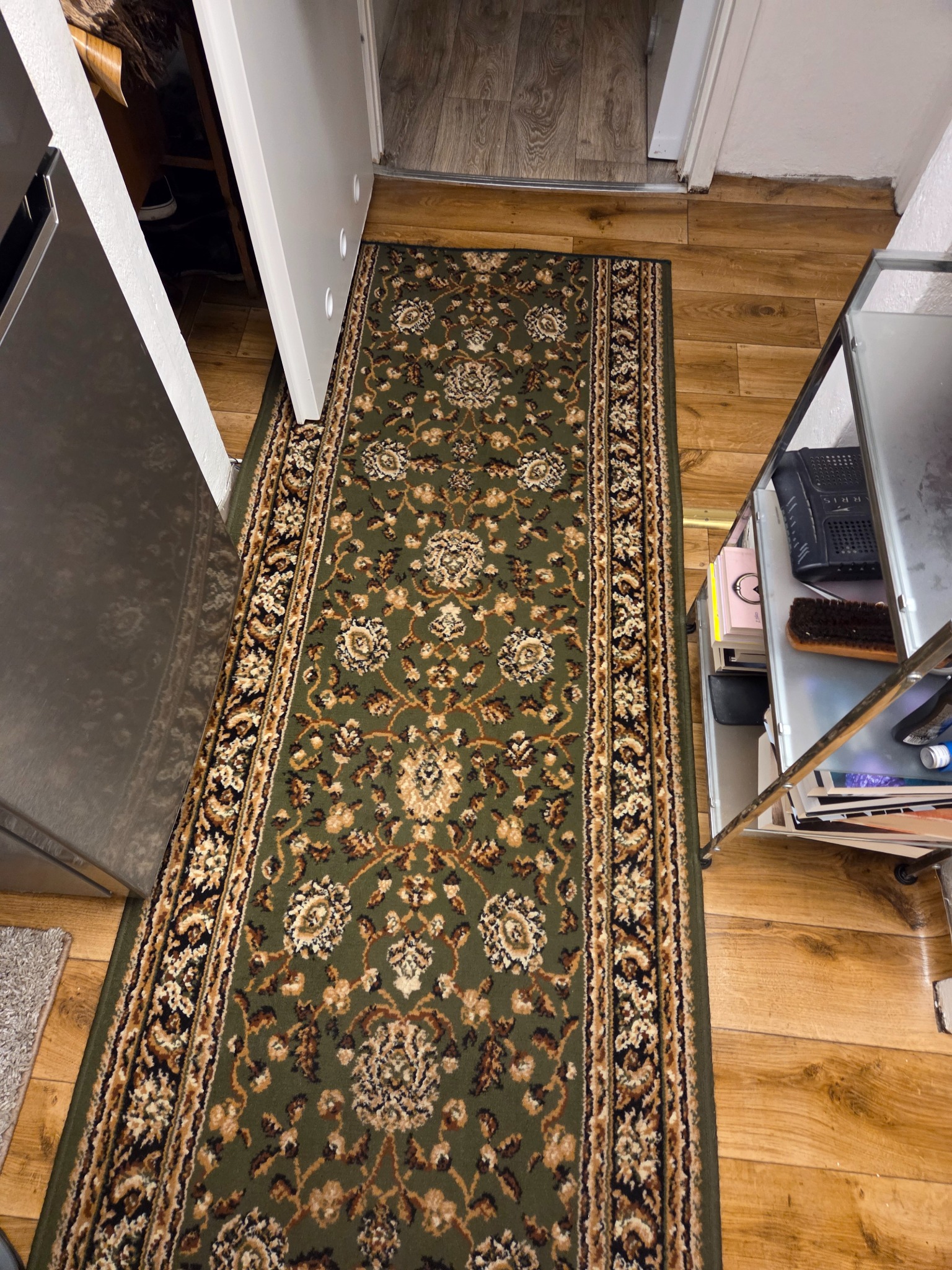Długi, zielony dywan z orientalnym wzorem leży na drewnianej podłodze w wąskim przejściu, obok metalowego regału z akcesoriami do czyszczenia.