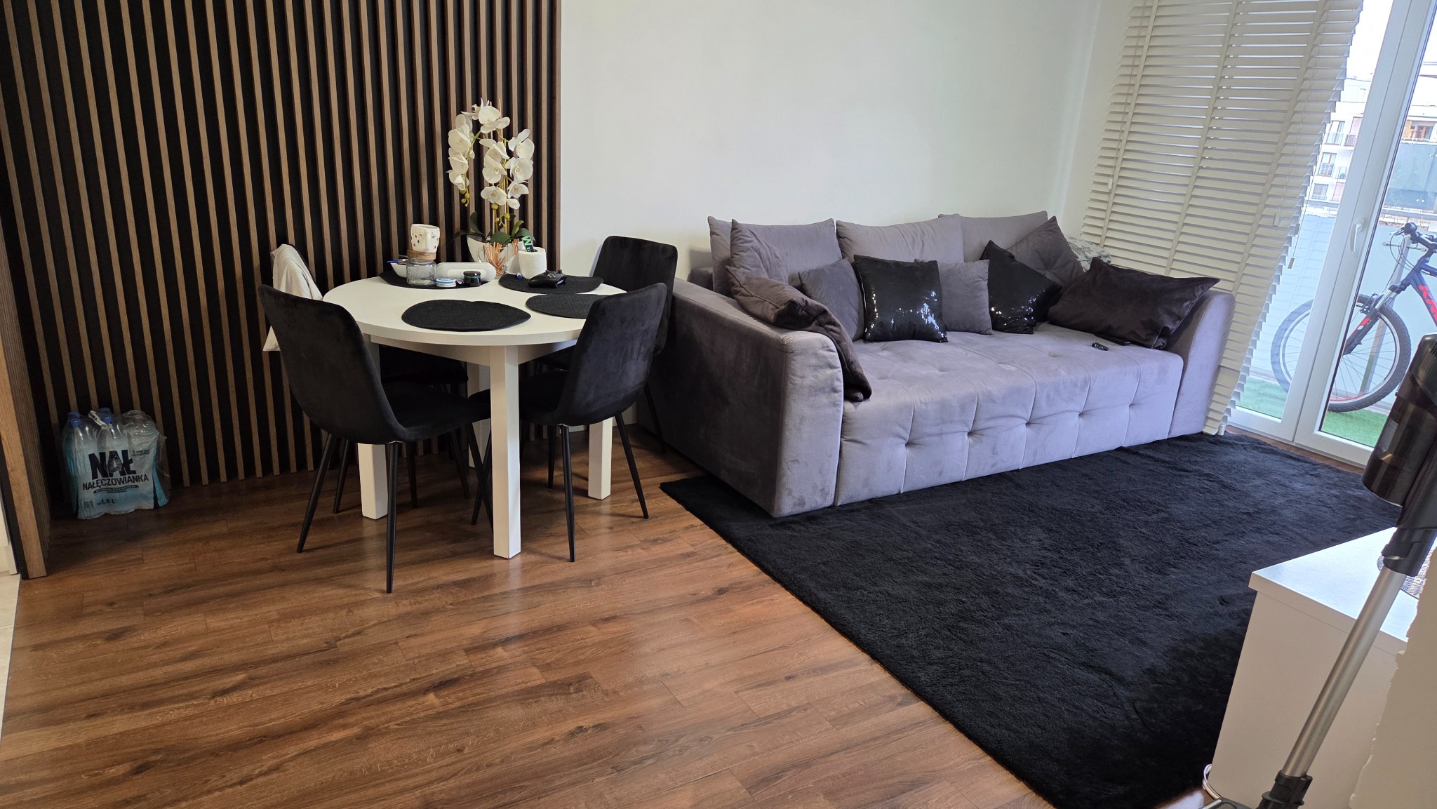 Wnętrze pokoju dziennego z szarą sofą, czarnym dywanem i stołem z krzesłami. Widoczne elementy dekoracyjne i butelki wody mineralnej przy ścianie.