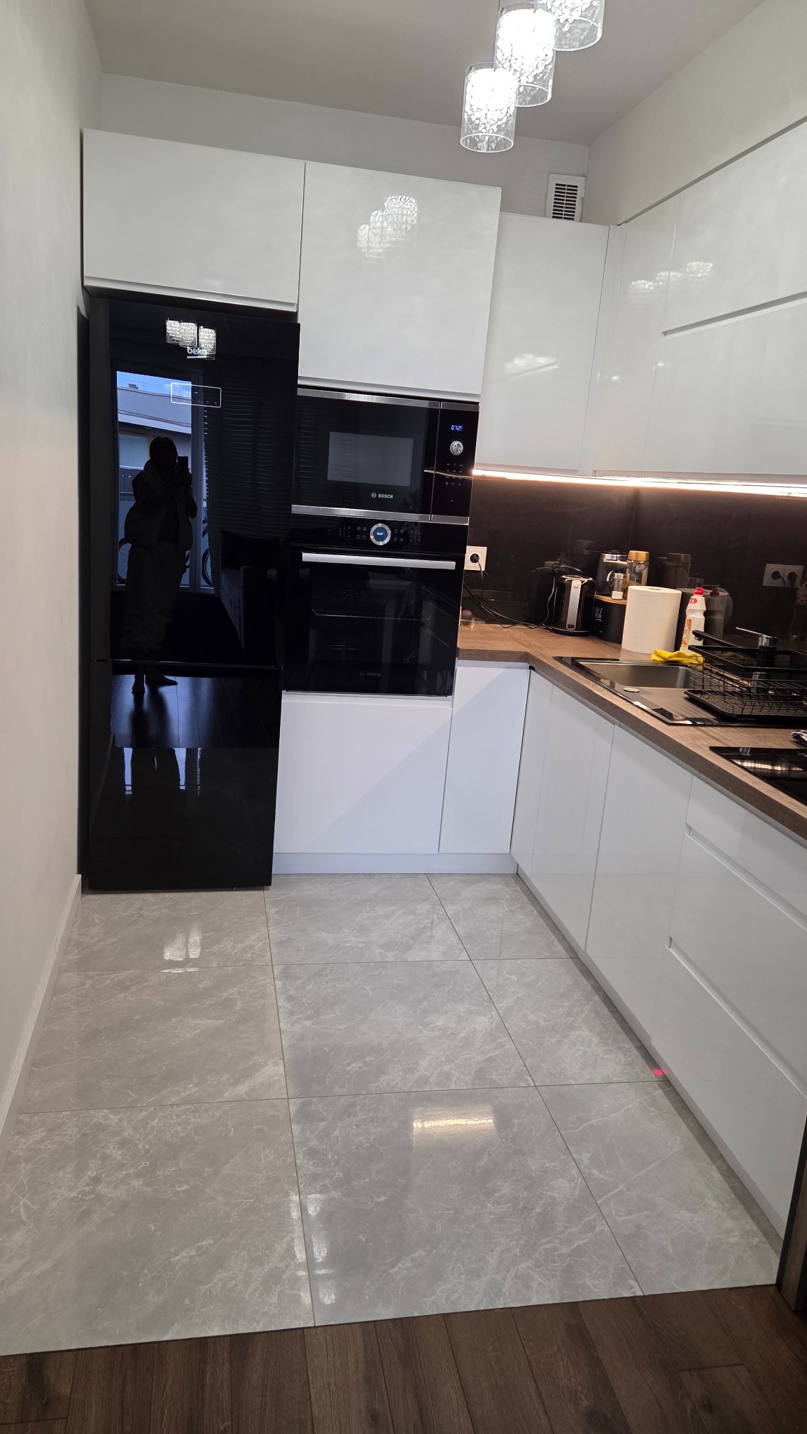 Nowoczesna kuchnia z białymi szafkami, czarnym sprzętem AGD marki Bosch i marmurową podłogą. Widoczne oświetlenie podszafkowe i designerskie lampy sufitowe.