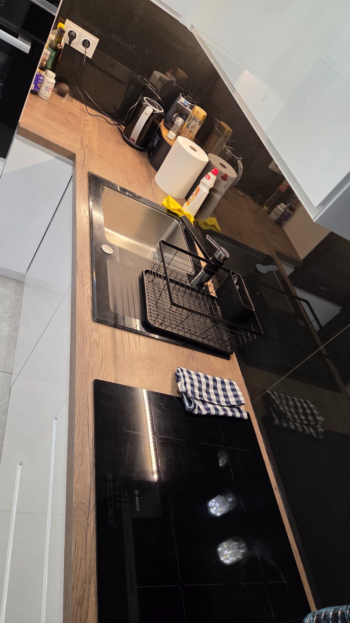 Błyszcząca, czarna płyta indukcyjna i zlewozmywak ze suszarką w nowoczesnej kuchni. Na blacie płyn do naczyń i ściereczka. Widoczne odbicia w blacie.