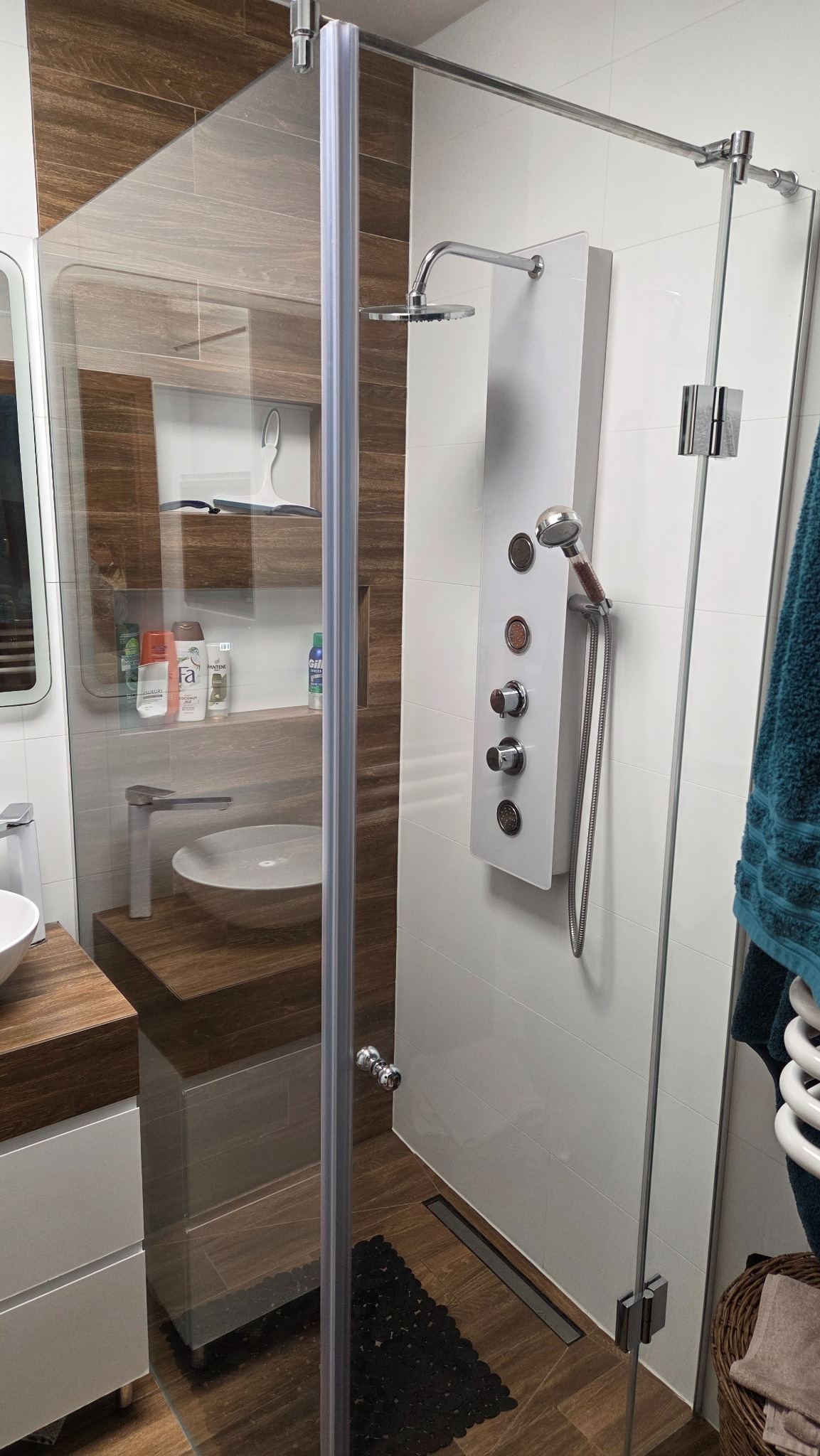 Nowoczesna kabina prysznicowa z panelem hydromasażu, szklaną ścianą i drewnianymi akcentami. Widoczna umywalka i półki z kosmetykami. Minimalistyczny design łazienki.