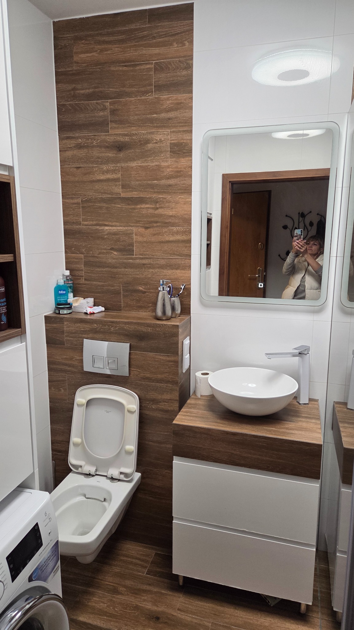 Nowoczesna łazienka po remoncie: drewnopodobne płytki, biała ceramika, umywalka nablatowa. W lustrze odbicie osoby robiącej zdjęcie.