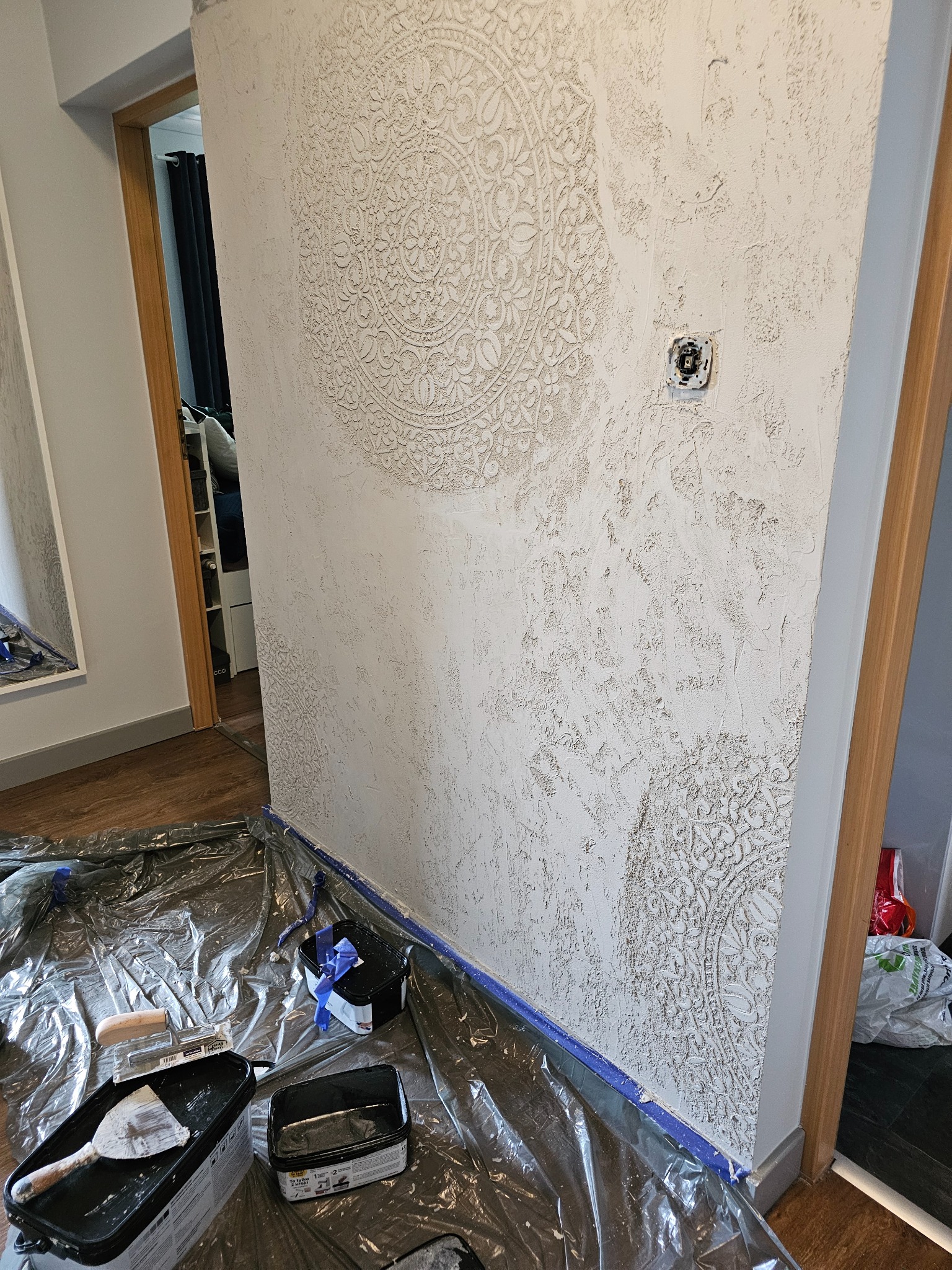 Ściana z dekoracyjnym tynkiem imitującym beton z ornamentami, zabezpieczona folią i taśmą malarską, widoczne narzędzia i puszki z farbą.