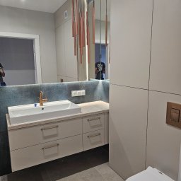 Front Meble Poznań - Nowoczesna łazienka z beżowymi szafkami pod umywalką, złotą armaturą i podłużnymi lampami wiszącymi w kolorze miedzi, widok na toaletę z boku