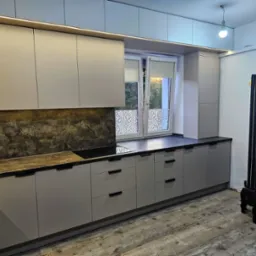 Szare, matowe szafki kuchenne z ciemnym blatem i podświetleniem LED, widok na okno z roletami i mały czarny pies w rogu pomieszczenia.