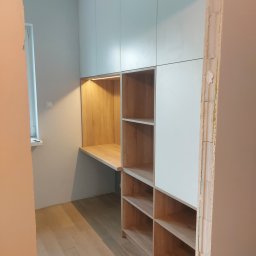 Front Meble Poznań - Wnęka z biurkiem i półkami z jasnego drewna, zintegrowana z szafą o białych frontach, oświetlona listwą LED, widok na surową ścianę i podłogę z desek.