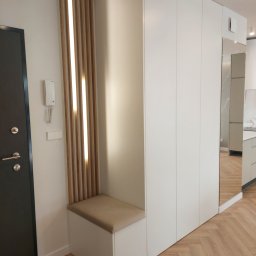 Front Meble Poznań - Nowoczesna szafa wnękowa z lustrem i siedziskiem, drewniane lamele z oświetleniem, minimalistyczny design, jasne kolory, widoczny fragment kuchni w odbiciu lustra.