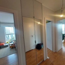 Front Meble Poznań - Biała szafa wnękowa z lustrzanymi drzwiami przesuwnymi, odbijająca wnętrze pokoju z białymi drzwiami i podłogą z paneli.