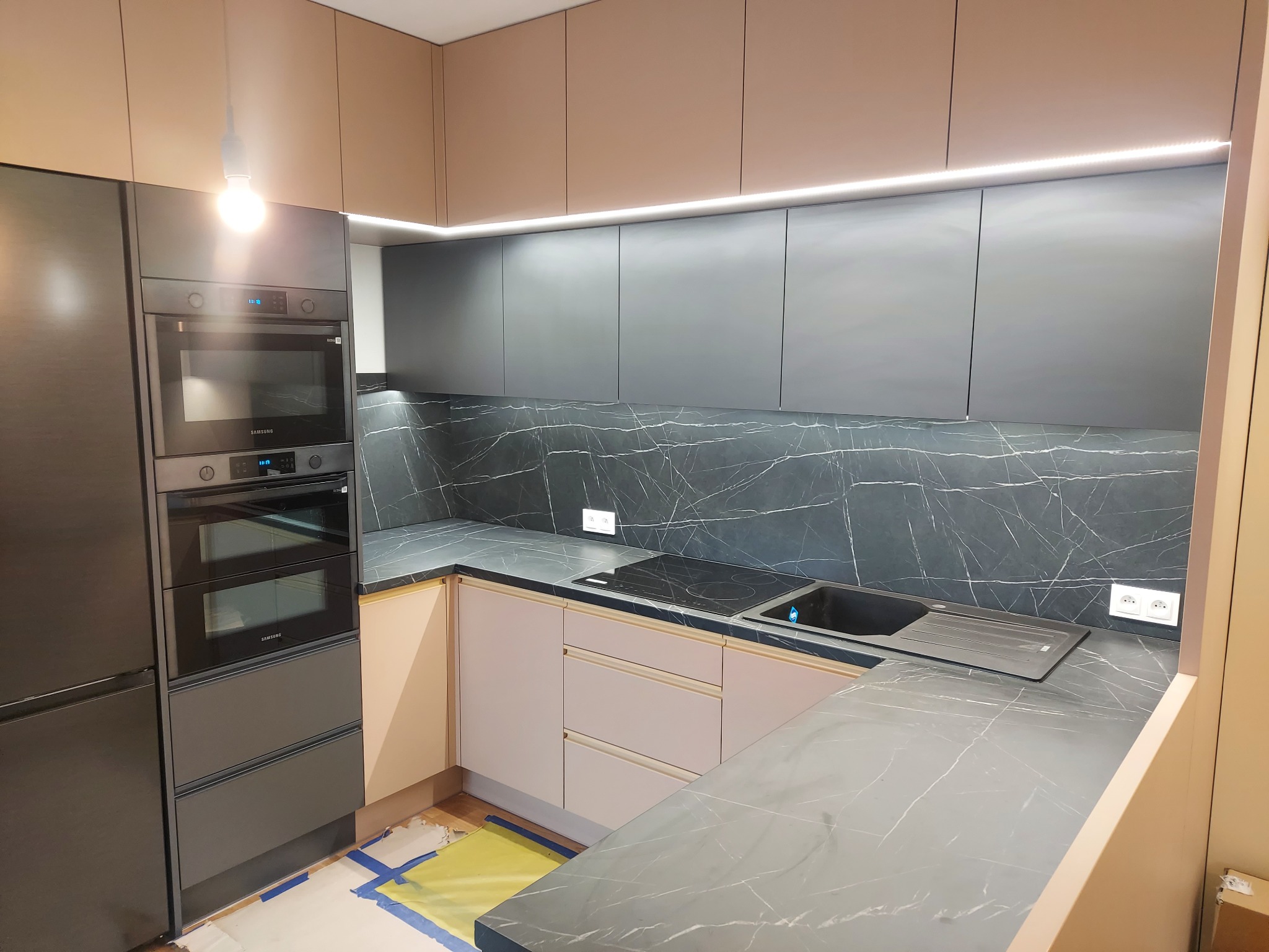 Nowoczesna kuchnia z szarymi szafkami górnymi i dolnymi w odcieniach beżu, marmurowy backsplash i blat, wbudowany piekarnik Samsung, podświetlenie LED pod szafkami górnymi, zabezpieczona podłoga...