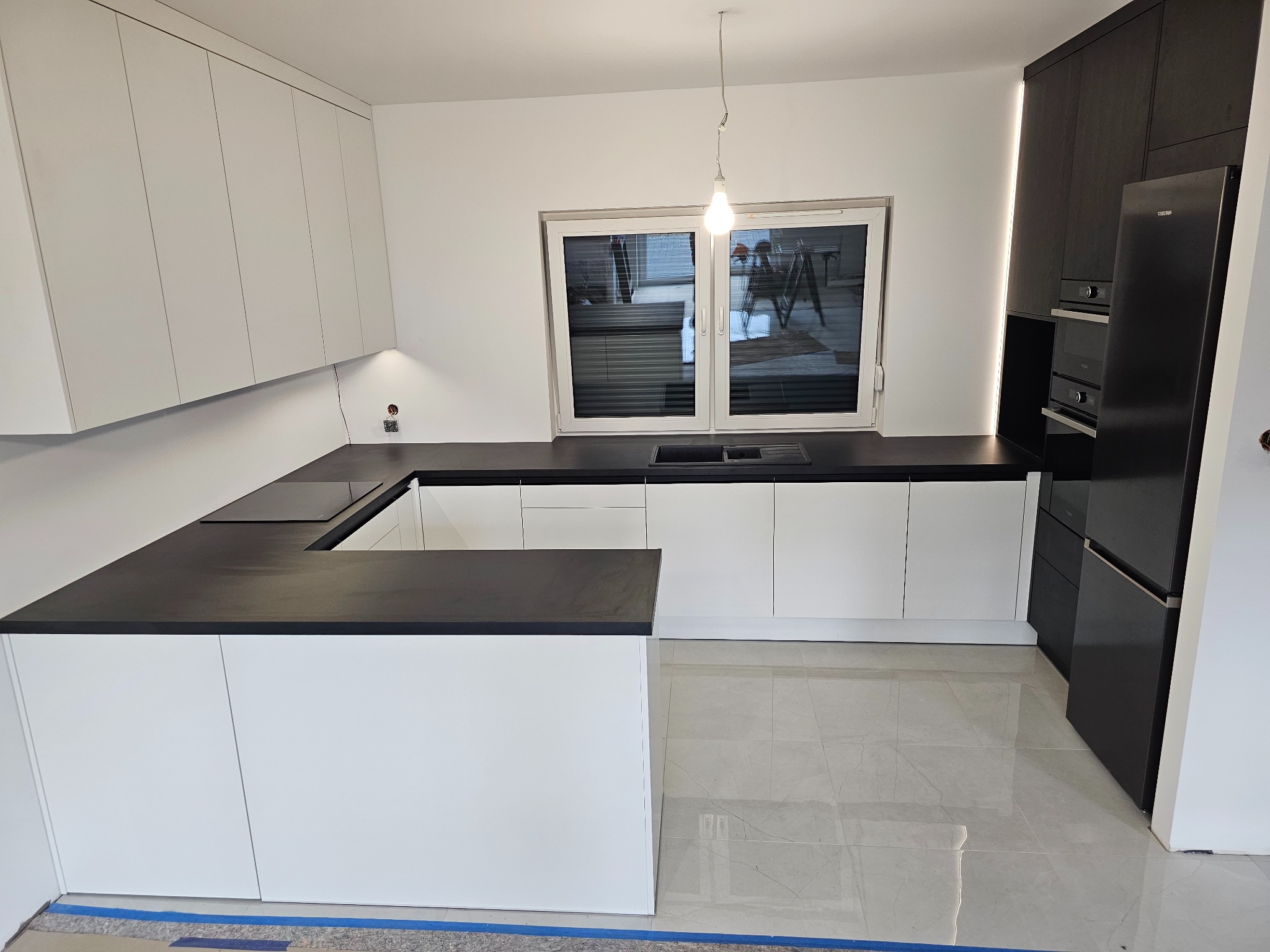 Nowoczesna kuchnia z białymi frontami szafek i czarnym blatem, oświetlenie punktowe pod szafkami wiszącymi, okno z widokiem na zewnątrz, płytki podłogowe imitujące marmur, zabudowa kuchenna...