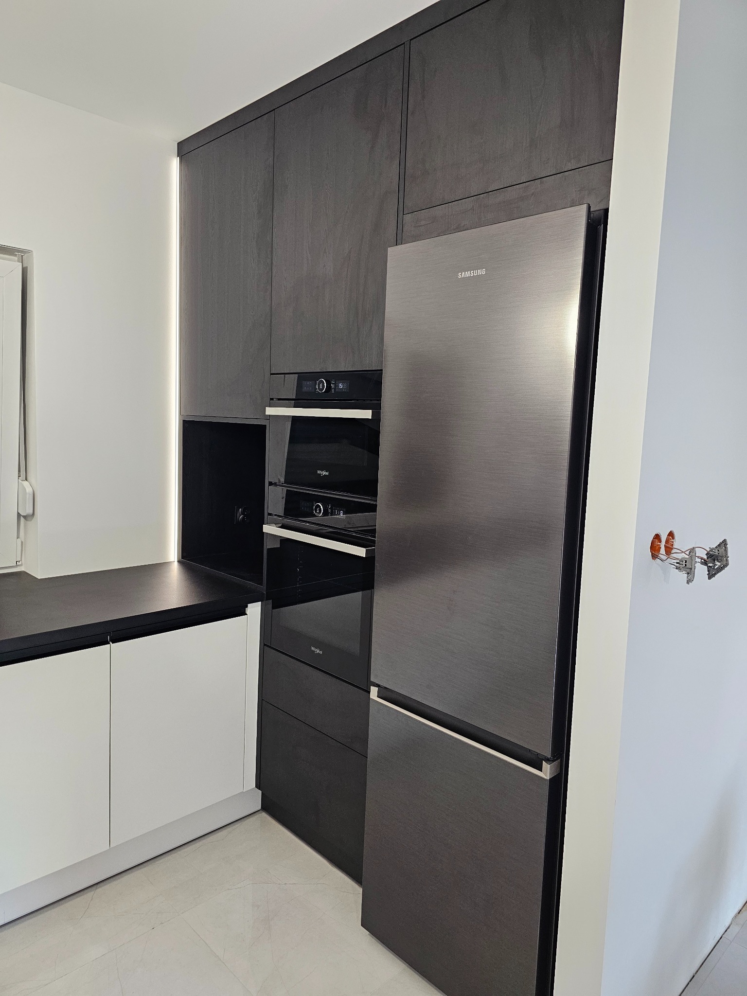 Nowoczesna kuchnia z zabudową w kolorze ciemnego drewna, zintegrowana z sprzętem AGD: lodówka Samsung, piekarnik i mikrofalówka Whirlpool, oświetlenie LED pod szafkami, białe dolne szafki...