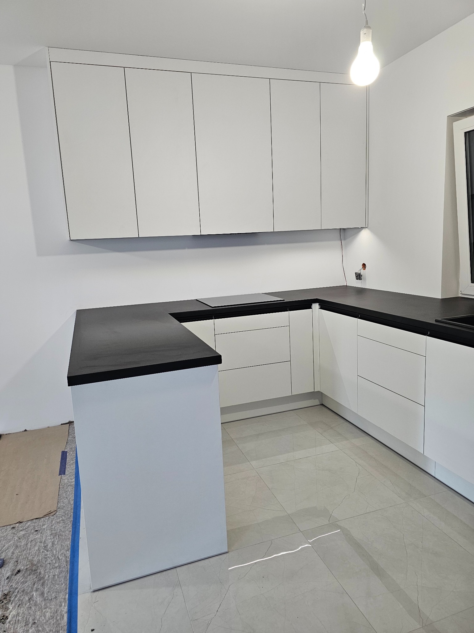 Nowoczesna, biała kuchnia w kształcie litery U z czarnym blatem, widoczne niedokończone prace wykończeniowe na podłodze.