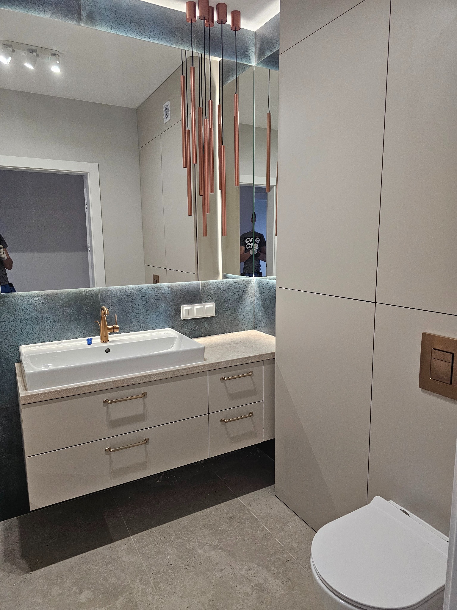 Nowoczesna łazienka z beżowymi szafkami pod umywalką, złotą armaturą i podłużnymi lampami wiszącymi w kolorze miedzi, widok na toaletę z boku